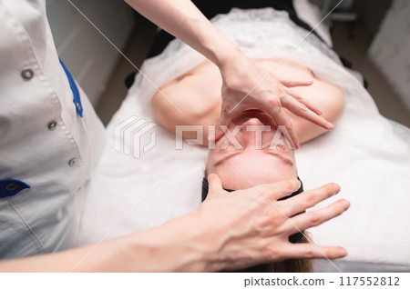 Facial massage beauty treatment. Close-up of a young woman face lying on back, getting face lifting massage, pinch and roll technique Facial massage beauty treatment. Close-up of a young woman face lying on back, getting face lifting massage, pinch and roll technique 117552812