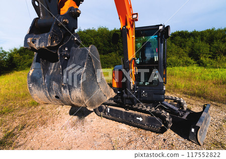 Excavator against the background of a green field and trees. Wide angle shooting Excavator against the background of a green field and trees. Wide angle shooting 117552822