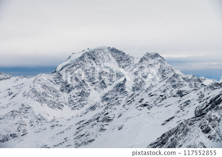 Picturesque view of mountain range with snow capped rocks under blue sky with white big cloud hanging over it in winter season Picturesque view of mountain range with snow capped rocks under blue sky with white big cloud hanging over it in winter season 117552855