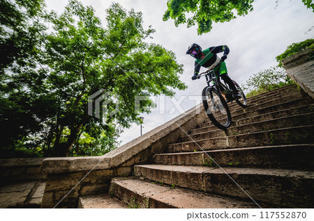 Male freerider on a bicycle quickly descends the city stairs. High-speed descent on a mountain bike down an old staircase among trees, panoramic view Male freerider on a bicycle quickly descends the city stairs. High-speed descent on a mountain bike down an old staircase among trees, panoramic view 117552870