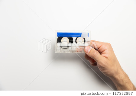 Close-up of a man's hand holding a blue cassette tape on a white background. Concept of music and sound, entertainment and nostalgia Close-up of a man's hand holding a blue cassette tape on a white background. Concept of music and sound, entertainment and nostalgia 117552897