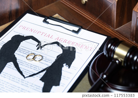 A clipboard holding a prenuptial agreement with a cut-out silhouette of a couple and wedding rings, next to a judge's gavel. A clipboard holding a prenuptial agreement with a cut-out silhouette of a couple and wedding rings, next to a judge's gavel. 117553383