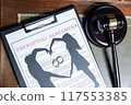 A clipboard holding a prenuptial agreement with a cut-out silhouette of a couple and wedding rings, next to a judge's gavel. 117553385