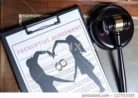 A clipboard holding a prenuptial agreement with a cut-out silhouette of a couple and wedding rings, next to a judge's gavel. 117553385