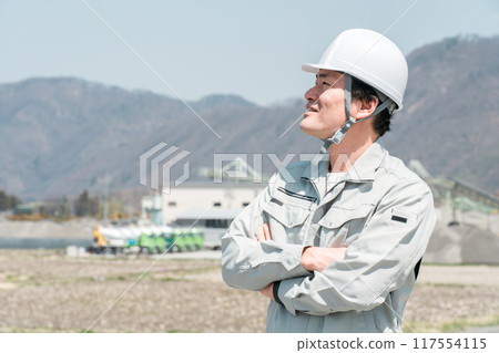 Asian male worker wearing work clothes and a helmet with his arms folded Asian male worker wearing work clothes and a helmet with his arms folded 117554115