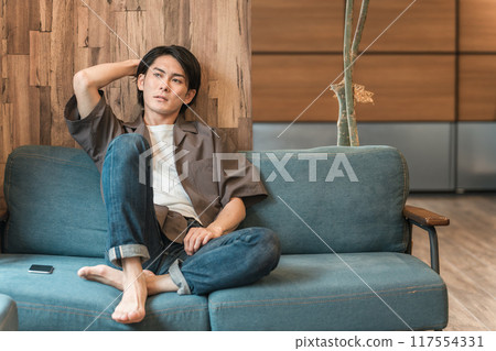 一個年輕的亞洲男子在客廳裡度過無聊的時光（無聊、沮喪、昏昏欲睡、遲鈍、疲倦、沮喪） 117554331