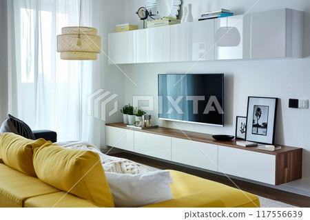 A bright and modern living room with a yellow sofa, white cabinets, and a large television 117556639