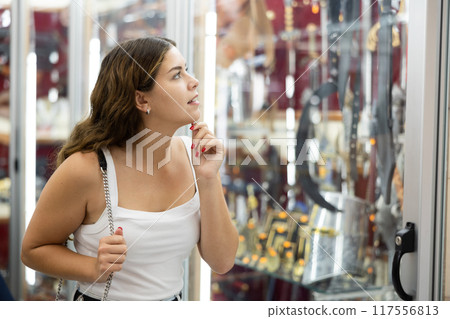Cheerful young girl viewing at forged swords in souvenirs shop 117556813