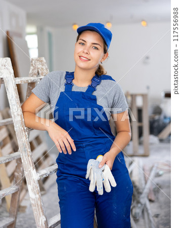 Portrait of positive builder woman in blue overalls next to stepladder Portrait of positive builder woman in blue overalls next to stepladder 117556988