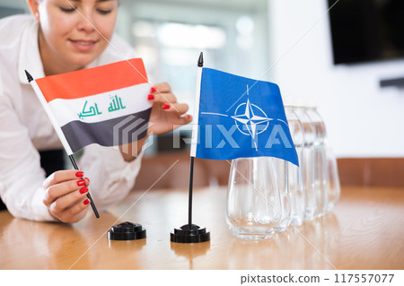 Woman secretary prepares an office for negotiations - she places flags of NATO (OTAN) and Iraq table Woman secretary prepares an office for negotiations - she places flags of NATO (OTAN) and Iraq table 117557077