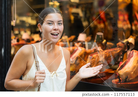 Passerby young woman examines smoked whole meat pork leg in shop window. 117557113