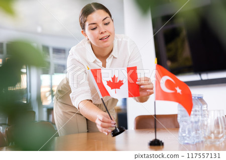 Positive young woman putting flag of Canada on the table with flag of Turkey in conference room 117557131