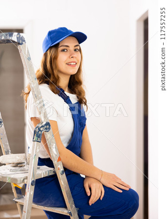 Portrait of young woman builder sitting on stepladder in apartment Portrait of young woman builder sitting on stepladder in apartment 117557255