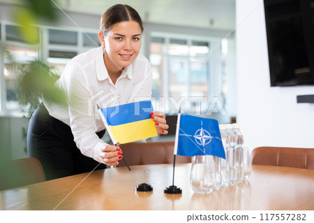 Businesswoman arranging the flags of NATO (OTAN) and Ukrain for presentation and negotiations Businesswoman arranging the flags of NATO (OTAN) and Ukrain for presentation and negotiations 117557282
