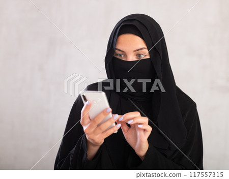 young positive girl with her face covered by burka holds phone in her hands and surf Internet 117557315