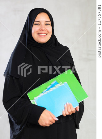 positive smiling girl in traditional black hijab stands and holds flock of colorful notebooks. positive smiling girl in traditional black hijab stands and holds flock of colorful notebooks. 117557391