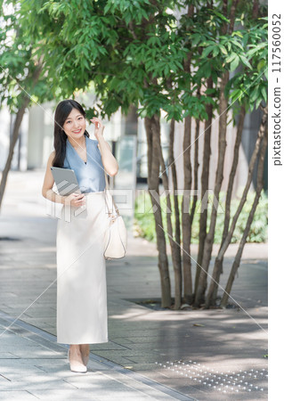 A businesswoman holding a tablet device in the shade of a tree in an office district A businesswoman holding a tablet device in the shade of a tree in an office district 117560052