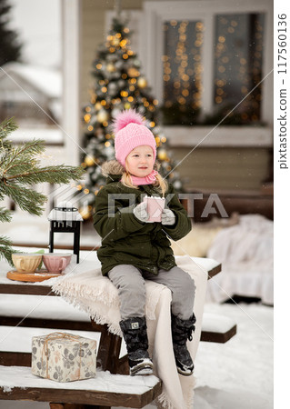 A little girl in winter clothes is sitting on the porch of a house decorated for the Christmas holidays. Christmas holidays for a child. A little girl in winter clothes is sitting on the porch of a house decorated for the Christmas holidays. Christmas holidays for a child. 117560136