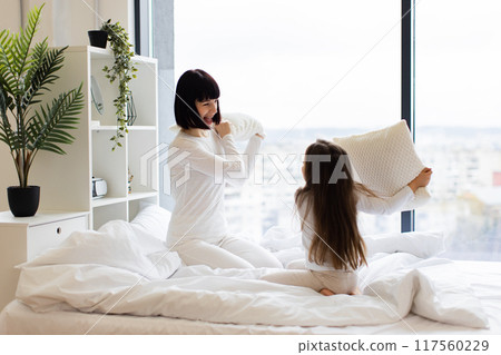 Mom and daughter pillow fight on cozy white bed Mom and daughter pillow fight on cozy white bed 117560229