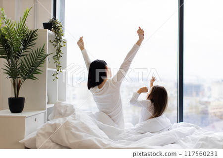 Mother and daughter stretching in bed by large window Mother and daughter stretching in bed by large window 117560231