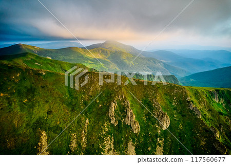 Breathtaking aerial view of green, rugged mountain peaks under dramatic sky. Scene captures contrast between vibrant landscape and moody clouds, with sunlight breaking through. Carpathians, Chornogora Breathtaking aerial view of green, rugged mountain peaks under dramatic sky. Scene captures contrast between vibrant landscape and moody clouds, with sunlight breaking through. Carpathians, Chornogora 117560677