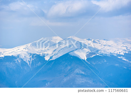 Snow-covered mountain peak under clear blue sky, with patches of snow on slopes and scattered trees. Serene winter landscape contrasts sharply with the dark, forested areas below. Blyznytsya, Ukraine 117560681