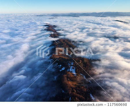 Breathtaking aerial view of mountain ridge piercing through sea of clouds. Sunlight illuminates patches of ridge. Distant mountain ranges create serene and expansive horizon. 117560698