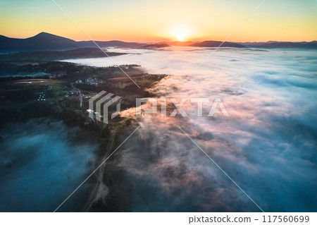 Stunning aerial view of sunrise over mountainous landscape, with thick fog blanketing valleys and hills. Sun's golden rays pierce through mist, casting serene glow over the entire scene. 117560699