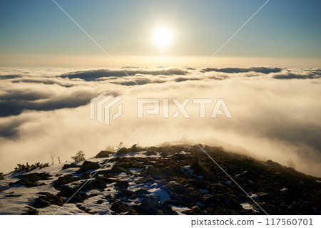 Sunrise over snow-covered mountain peak. Warm golden light of rising sun bathes clouds in soft hues of orange and pink. Snow and scattered rocks in foreground add texture and depth to scene. 117560701