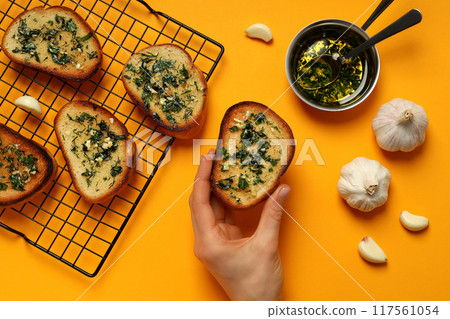 Slices of garlic bread on grid and in hand, garlic and bowl with oil on yellow background, top view 117561054