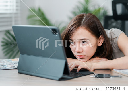 A young woman rests her head on her arms while working on a tablet in a home office, appearing tired and bored. A young woman rests her head on her arms while working on a tablet in a home office, appearing tired and bored. 117562144
