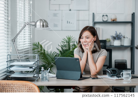 A young woman in a modern home office, excitedly working on a tablet, surrounded by natural light and greenery. A young woman in a modern home office, excitedly working on a tablet, surrounded by natural light and greenery. 117562145