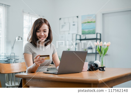 A young woman using a credit card to shop online with a laptop in a modern home office, surrounded by decor and natural light. A young woman using a credit card to shop online with a laptop in a modern home office, surrounded by decor and natural light. 117562148
