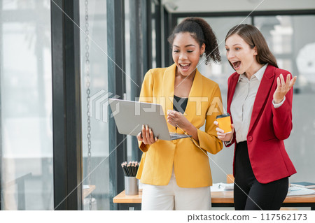Two excited businesswomen in professional attire working together on a laptop in a bright, modern office space. 117562173