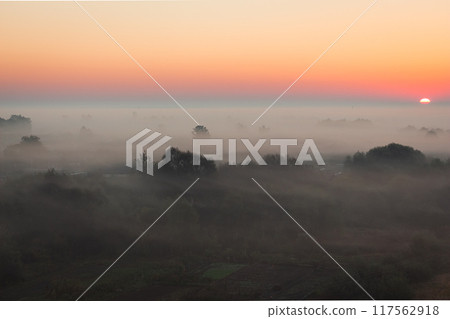 A serene dawn unfolds peacefully over a tranquil, misty landscape bathed in a vibrant horizon 117562918