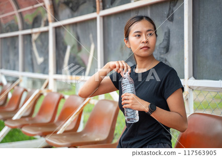 Young Asian female runner opening up the cap of her water bottle during her running break Young Asian female runner opening up the cap of her water bottle during her running break 117563905