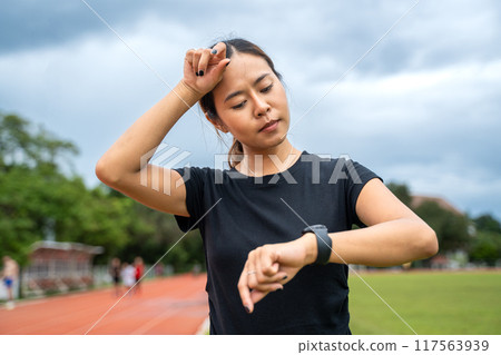 Young Asian female runner looking at her run time on her watch and wipe off her sweat from her forehead after her evening run 117563939