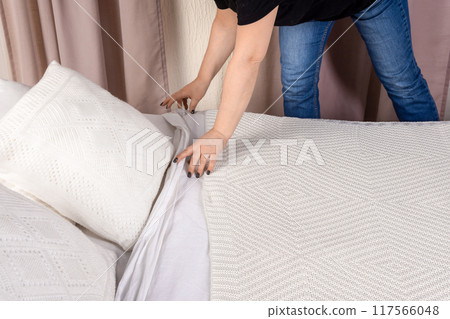 Young woman making the bed with white pillows and blanket in a cozy bedroom scene Young woman making the bed with white pillows and blanket in a cozy bedroom scene 117566048