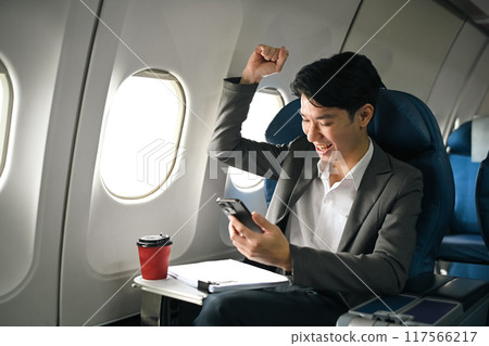 Excited young businessman looking at his smartphone and celebrating success in an airplane seat 117566217