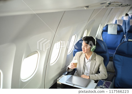 Pleasant young businessman with a cup of coffee listening to music in headphones sitting on plane during a flight Pleasant young businessman with a cup of coffee listening to music in headphones sitting on plane during a flight 117566223