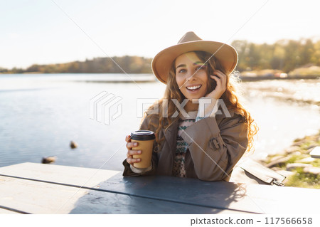 Smiling woman in hat on terrace with phone enjoys sunny autumn weather. Concept of relaxation, rest. Smiling woman in hat on terrace with phone enjoys sunny autumn weather. Concept of relaxation, rest. 117566658
