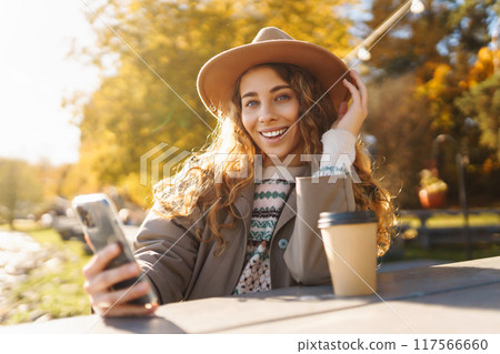 Smiling woman in hat on terrace with phone enjoys sunny autumn weather. Concept of relaxation, rest. Smiling woman in hat on terrace with phone enjoys sunny autumn weather. Concept of relaxation, rest. 117566660
