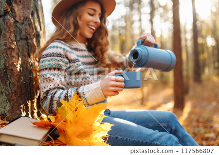 Woman traveler in hat sits near tree with thermos, enjoys autumn nature. Concept of nature. Woman traveler in hat sits near tree with thermos, enjoys autumn nature. Concept of nature. 117566807