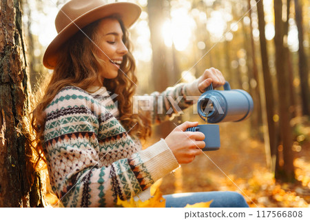Woman traveler in hat sits near tree with thermos, enjoys autumn nature. Concept of nature. Woman traveler in hat sits near tree with thermos, enjoys autumn nature. Concept of nature. 117566808