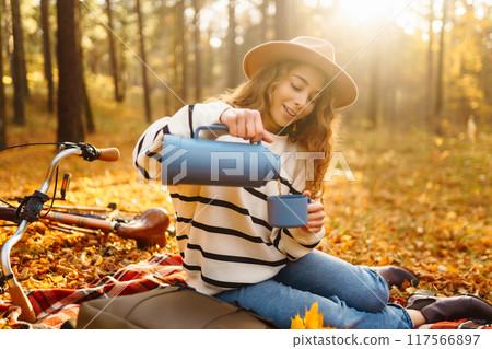 Hiker sitting on picnic blanket, drinking hot drink in autumn forest. Woman resting in nature. 117566897