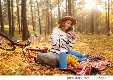 Hiker sitting on picnic blanket, drinking hot drink in autumn forest. Woman resting in nature. 117566898