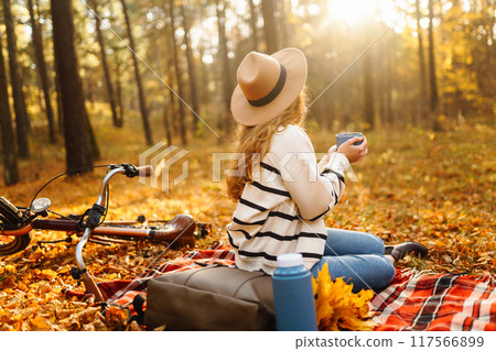 Hiker sitting on picnic blanket, drinking hot drink in autumn forest. Woman resting in nature. Hiker sitting on picnic blanket, drinking hot drink in autumn forest. Woman resting in nature. 117566899