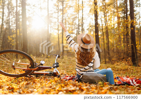 Hiker sitting on picnic blanket, drinking hot drink in autumn forest. Woman resting in nature. Hiker sitting on picnic blanket, drinking hot drink in autumn forest. Woman resting in nature. 117566900