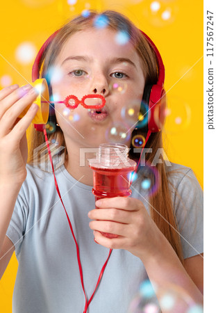 Cute girl with headphones blowing soap bubbles on yellow background 117567247