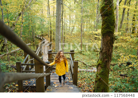 Woman in a yellow coat with backpack walks along wooden staircase, enjoying autumn scenery in forest Woman in a yellow coat with backpack walks along wooden staircase, enjoying autumn scenery in forest 117568448
